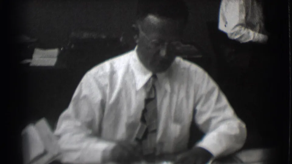 Adult man in office dress turns slightly at a desk with papers in 1959 Saint Louis home-movie footage.