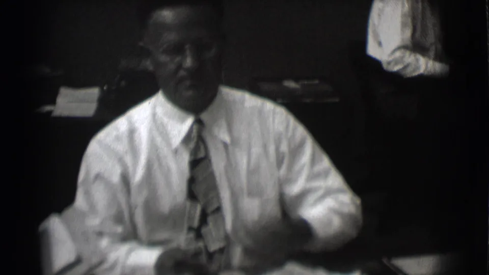 In 1959 Saint Louis footage, a man in glasses, white shirt, and tie sits at a desk in a dim office.