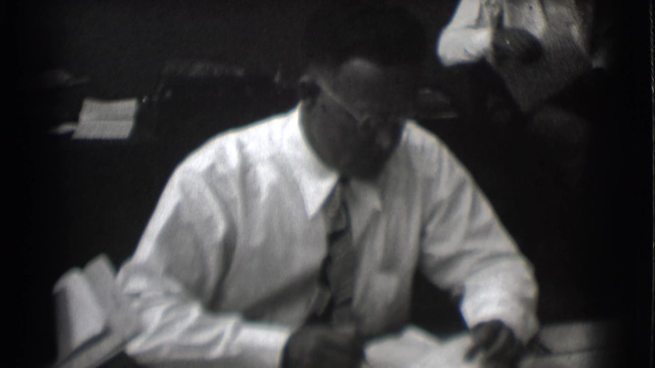 The businessman looks down toward paperwork at his desk in the 1959 Saint Louis office scene.