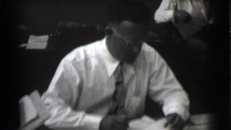The businessman looks down toward paperwork at his desk in the 1959 Saint Louis office scene.