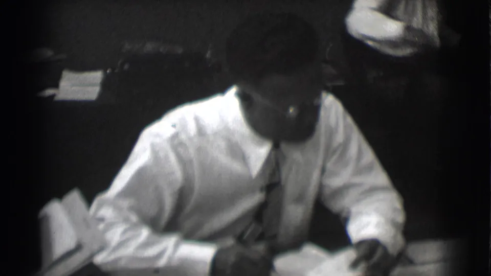 A man in a white shirt and tie works over papers at a desk in black-and-white 8mm office footage.