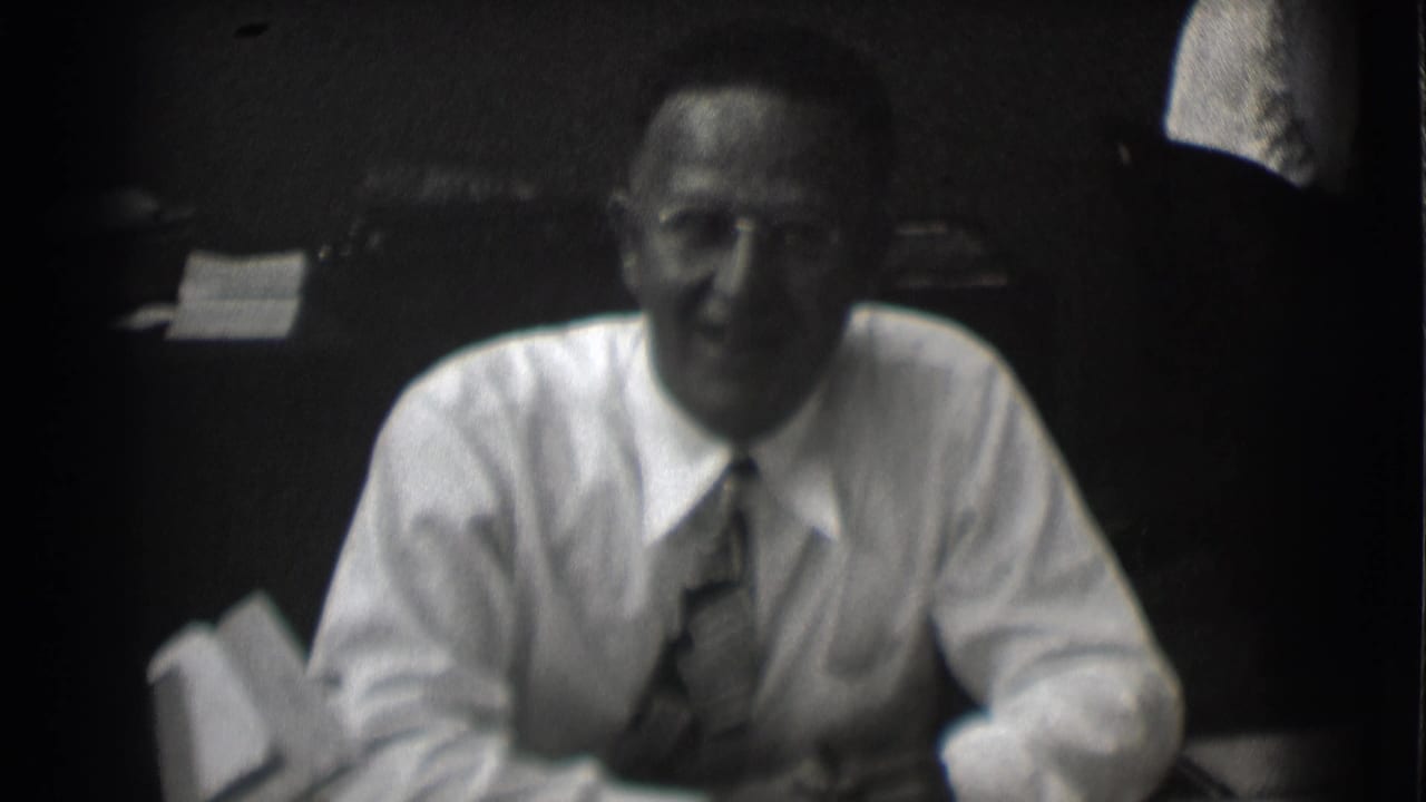 Man in a white shirt and tie seated at a desk in a dark office.