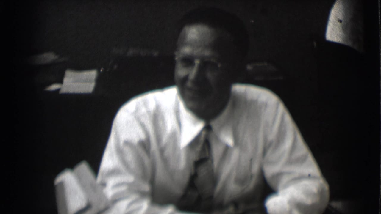 Smiling man in glasses sits at a desk with papers behind him.
