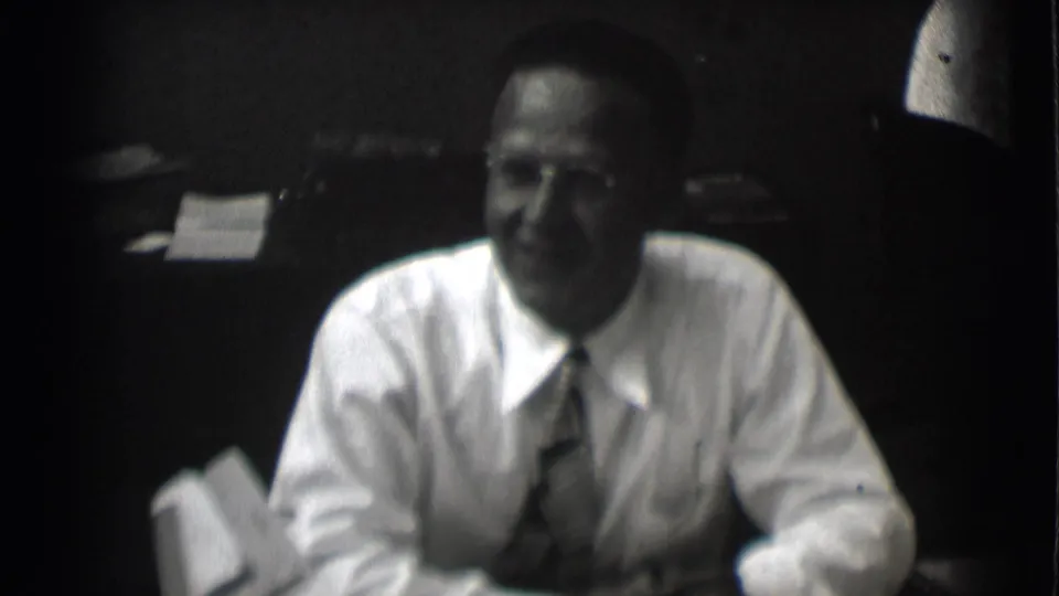 The man appears to smile while seated at a desk, with papers and a dark office background visible in the 1959 Saint Louis clip.