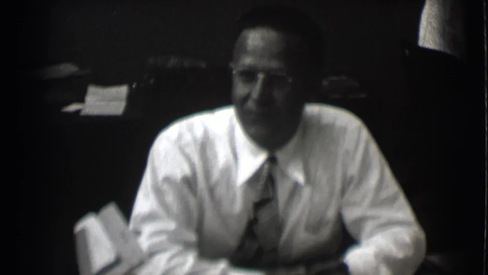 A man in a white dress shirt and tie leans forward at a desk in soft black-and-white 8mm footage from Saint Louis, 1959.