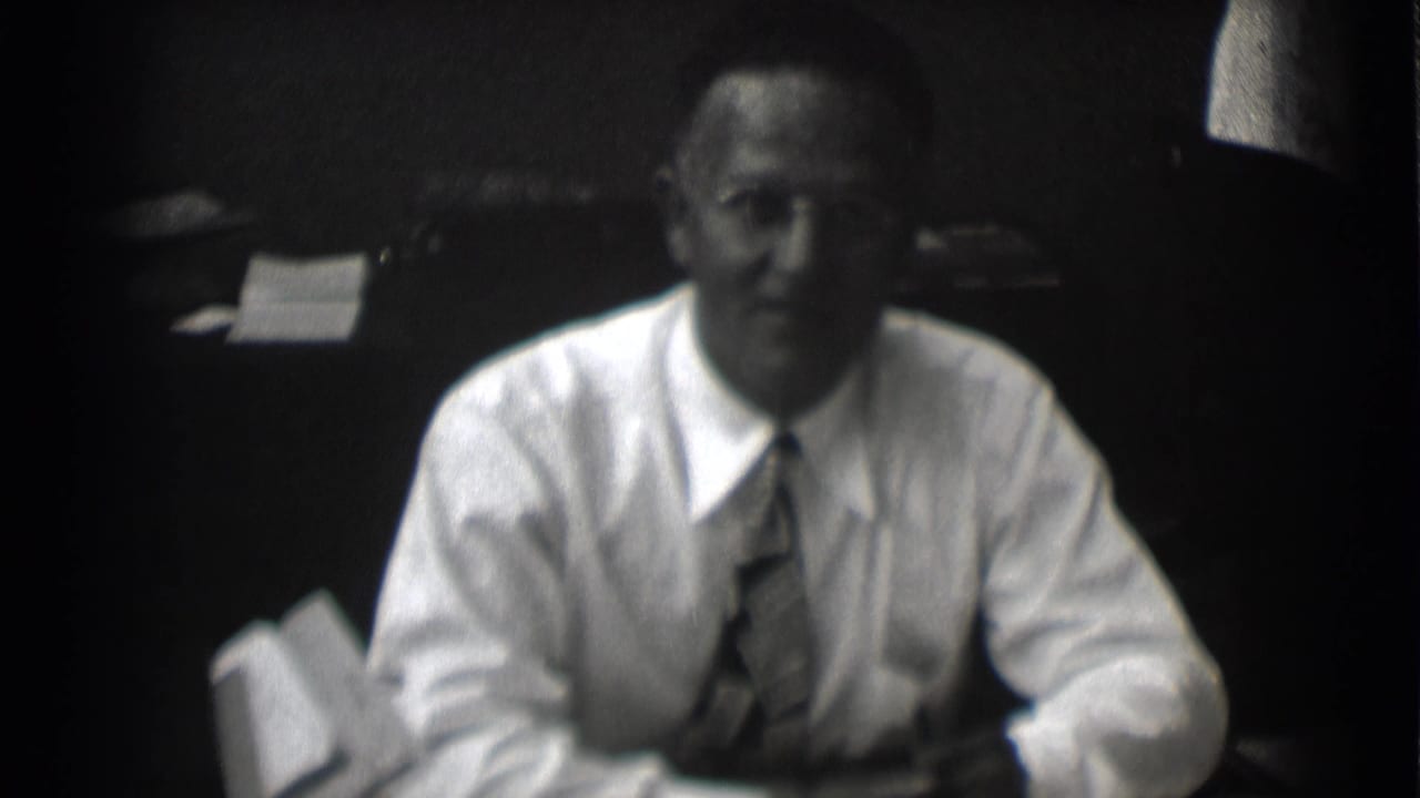 Man in a white shirt and tie sits at a desk facing the camera.