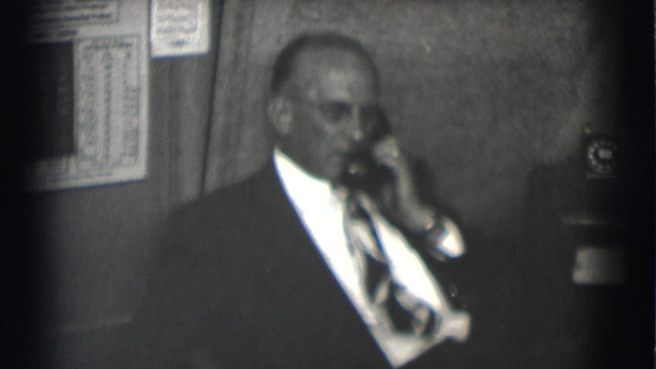 Suited adult man wearing glasses speaks on a rotary telephone inside an office setting in Saint Louis, Missouri.