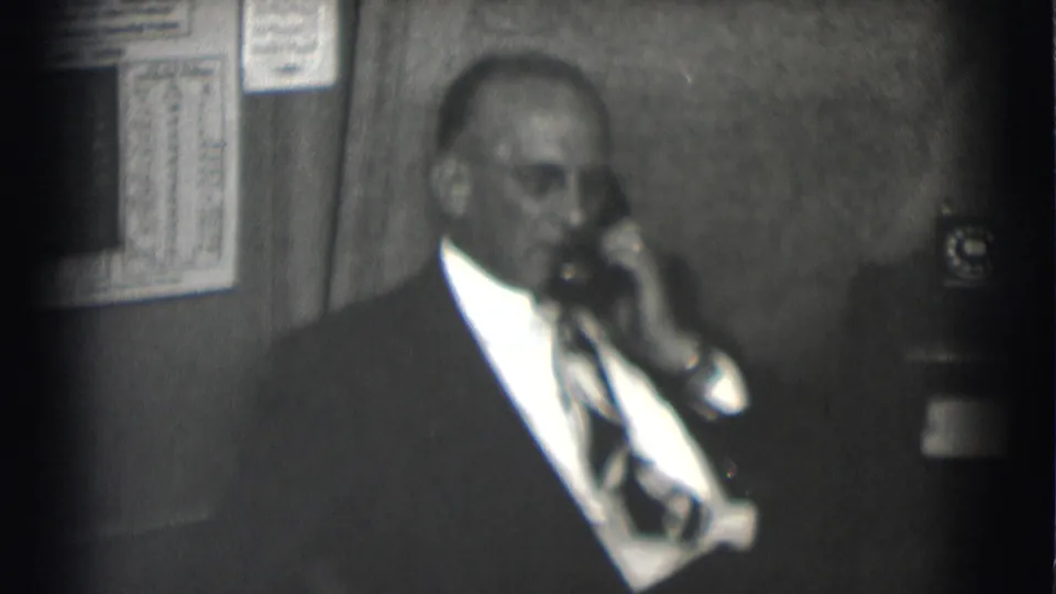 Suited adult man wearing glasses speaks on a rotary telephone inside an office setting in Saint Louis, Missouri.