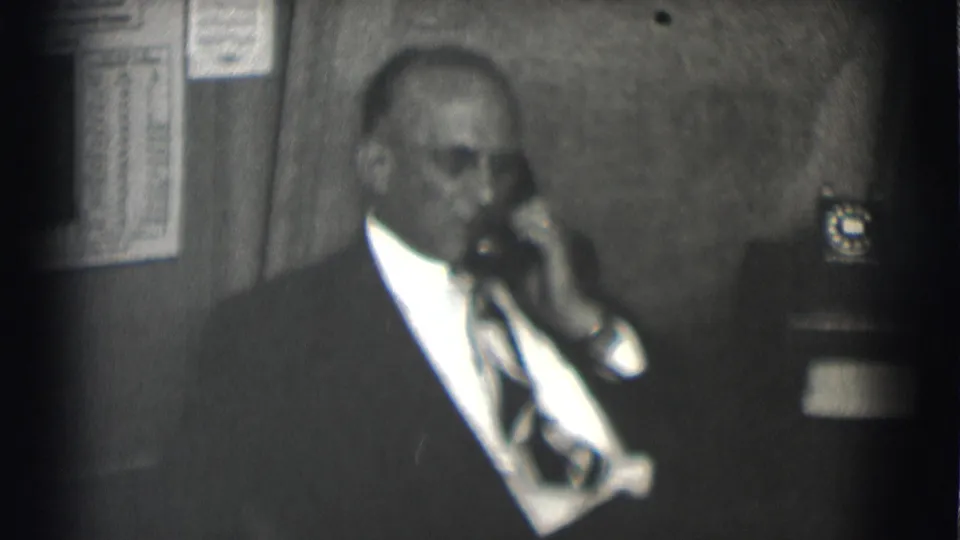 Adult man in a suit and tie remains on a rotary telephone in a softly focused 1959 office interior.