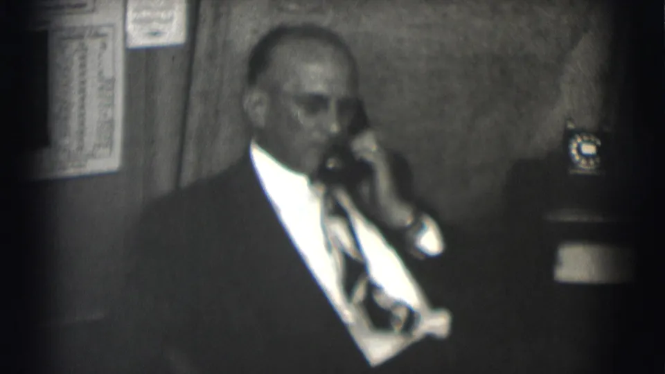 Suited man with glasses uses a rotary telephone while seated in an office-like room, captured on grainy 8mm film.