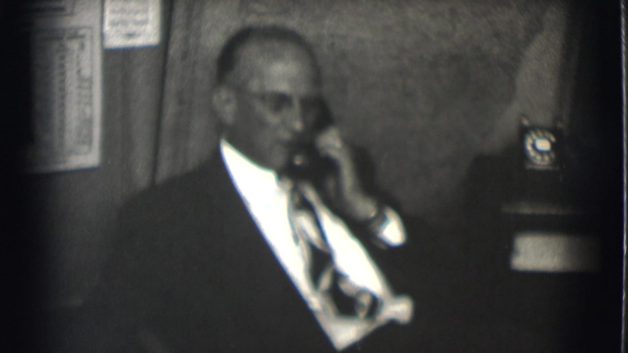 In Saint Louis 1959 footage, a suited man with glasses holds a rotary phone receiver beside a wall chart and desk phone.