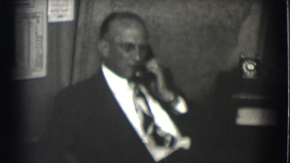 In Saint Louis 1959 footage, a suited man with glasses holds a rotary phone receiver beside a wall chart and desk phone.