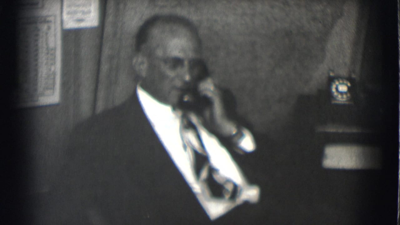 Suited man listens on a telephone with an office phone behind him.