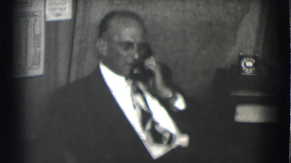 A man in a dark suit and tie listens on a telephone in the Saint Louis office interior, with another phone visible nearby.