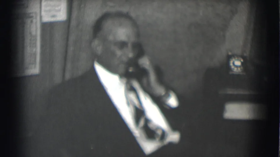 The 1959 8mm frame shows the suited man speaking into the phone, with a wall chart and spare desk phone in the background.