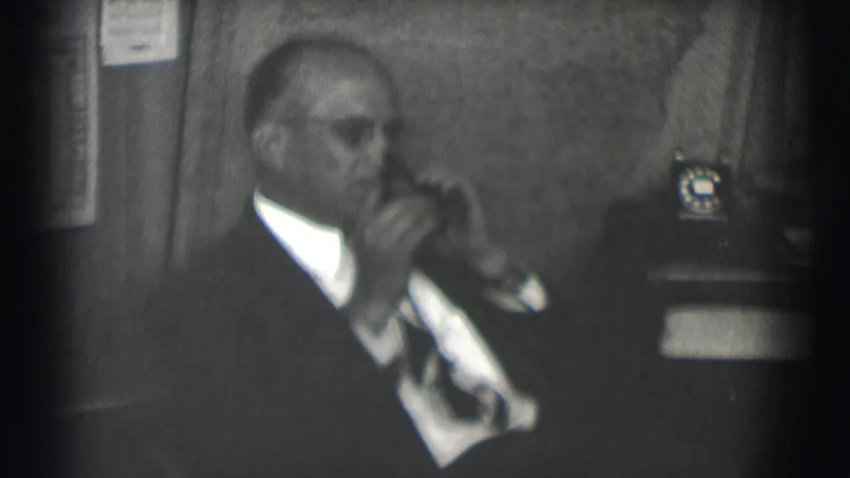 The man turns in profile while continuing the office phone call, with soft black-and-white 8mm blur and a desk phone behind him.