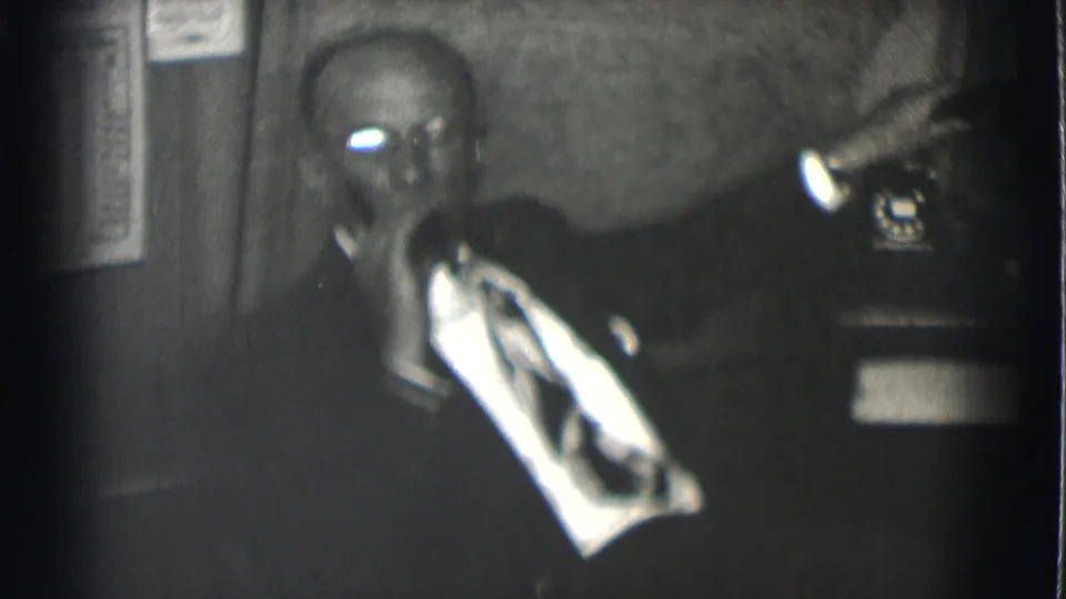In a dim Saint Louis office setting from 1959, a suited man sits at a desk with a telephone receiver near his face.