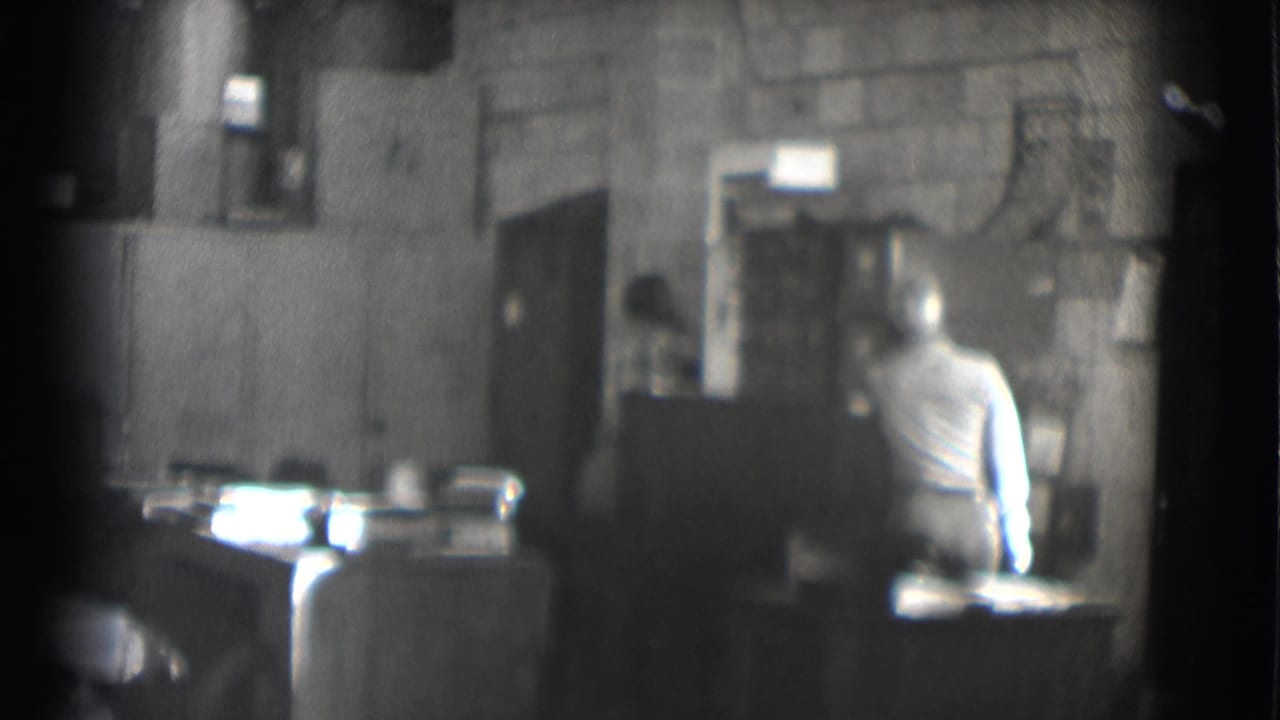 Man in a light shirt sits at a desk in a dim office with block walls.