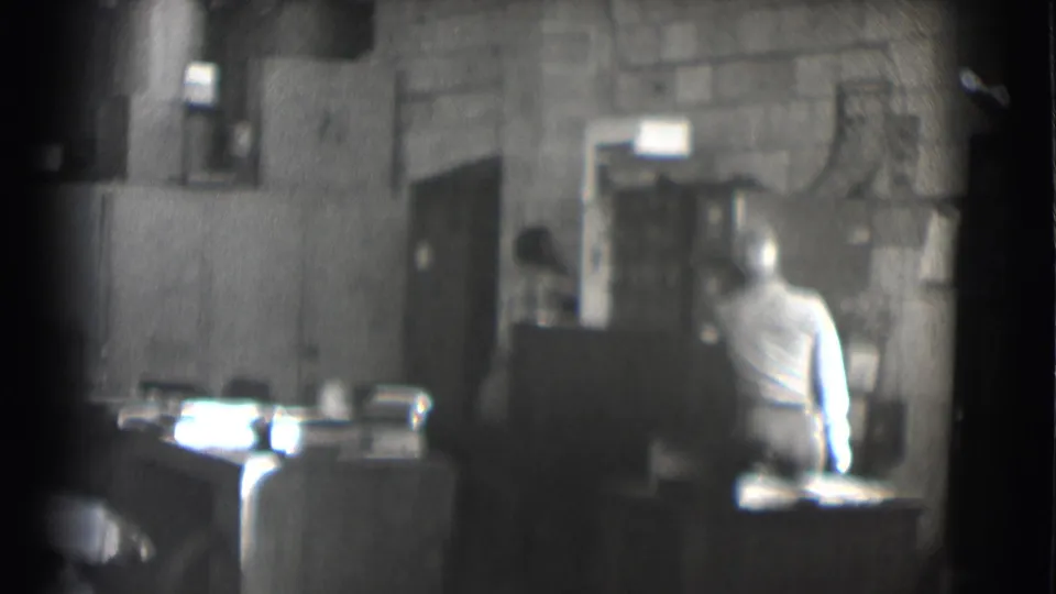 1959 Saint Louis 8mm frame showing a seated man in a light shirt facing a desk inside a dim office or workroom.