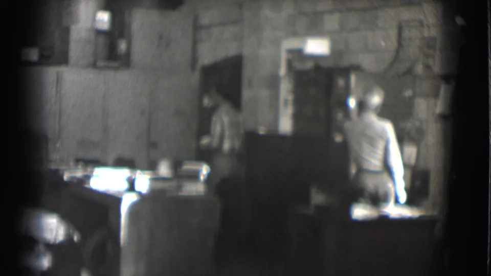 1959 Saint Louis office or workroom frame showing a seated man at a desk and another person partly visible by the doorway.