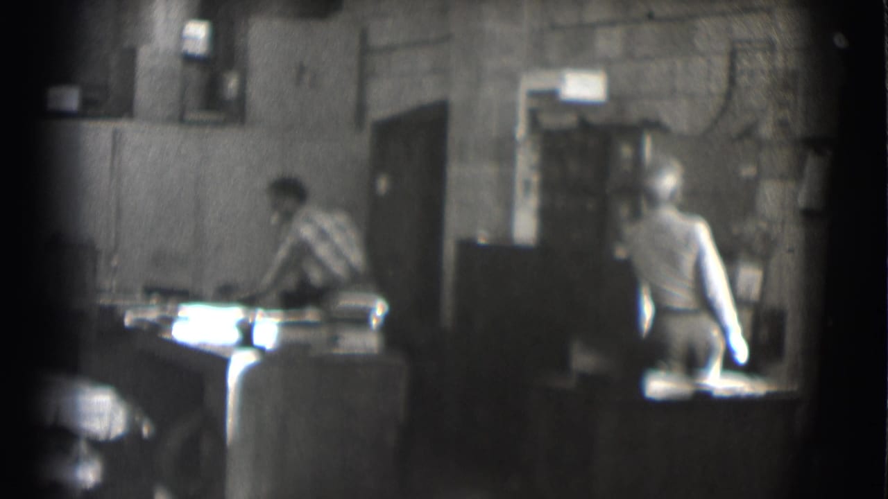 Two men in a dim workshop or office, one leaning over a desk while another stands nearby.