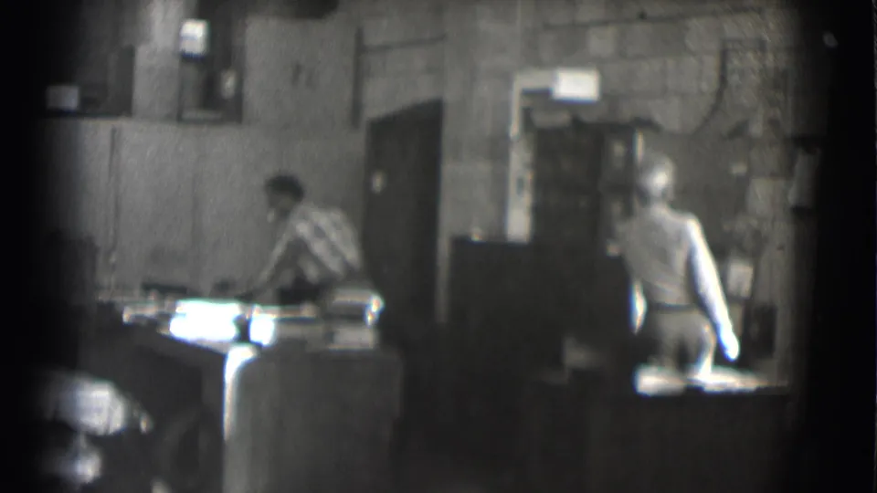 1959 Saint Louis 8mm frame showing two adult men in a dim interior, with one working at a table and another standing near cabinets.