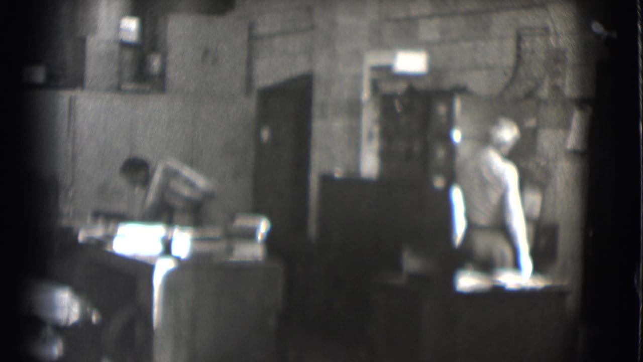 1959 Saint Louis workshop or office frame with one man leaning over papers and another standing near a cabinet or doorway.