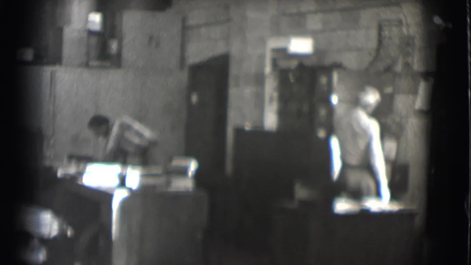 1959 Saint Louis workshop or office frame with one man leaning over papers and another standing near a cabinet or doorway.