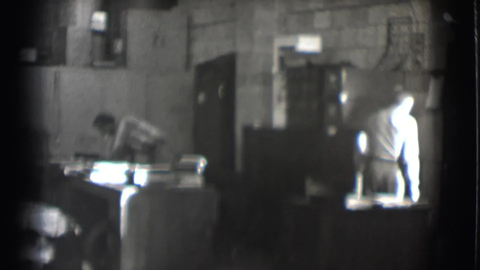 Grainy 1959 Saint Louis 8mm frame of two men in a workshop or office, with papers and furniture visible around them.