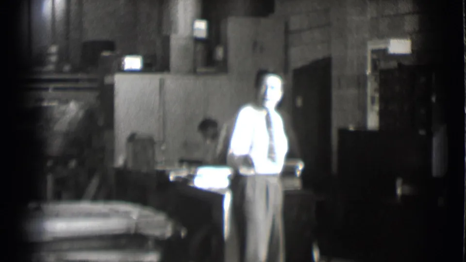 1959 Saint Louis 8mm frame showing a man in shirt and tie in the foreground with another man working at a table behind him.