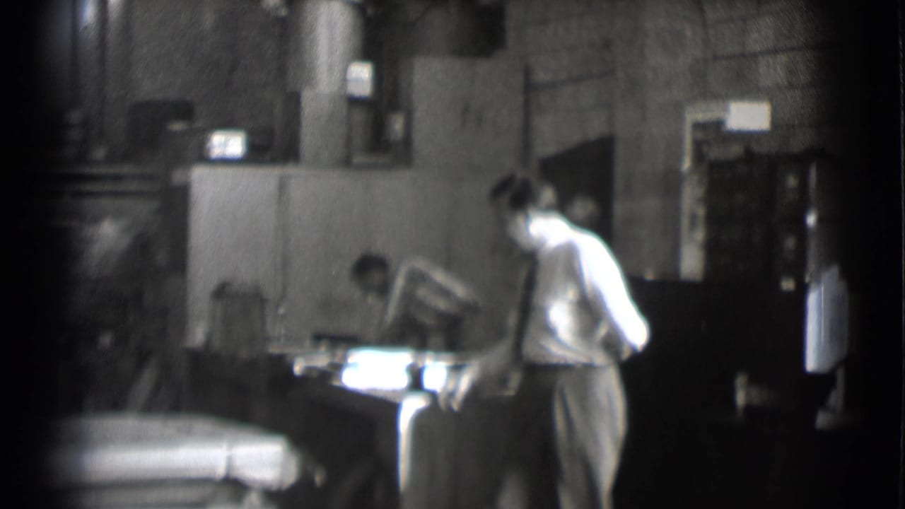 Dim newsroom office with a man in a light shirt and tie standing near a desk.