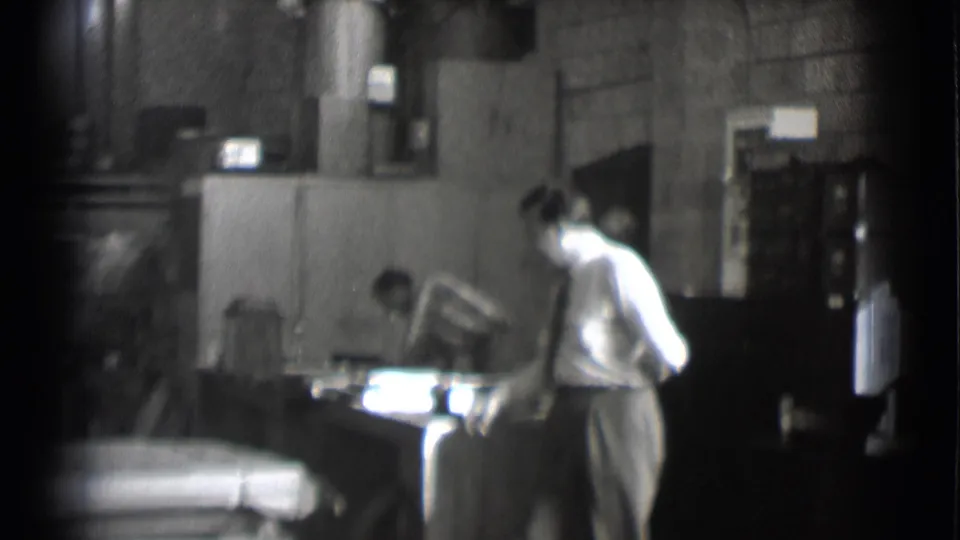 1959 Saint Louis newsroom interior with a man in shirt and tie beside a desk in a grainy black-and-white 8mm frame.
