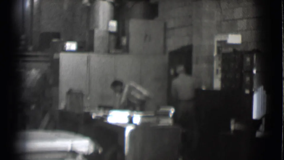 A worker leans over a desk or console inside the 1959 Saint Louis newsroom, with equipment and wall details behind him.