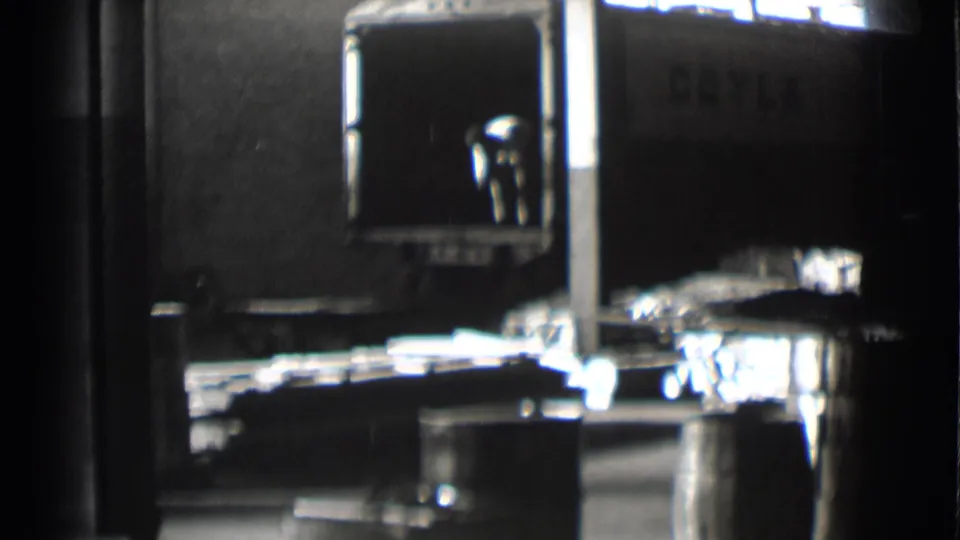 Saint Louis factory footage shows a worker beside the loading area, with metal sheets, a truck side, and harsh overhead light.