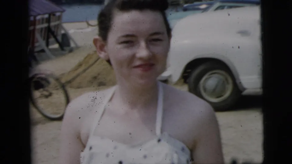 In Camden, New Jersey, 1961 footage, a young woman in a white patterned swimsuit smiles toward the camera near mid-century parked cars.