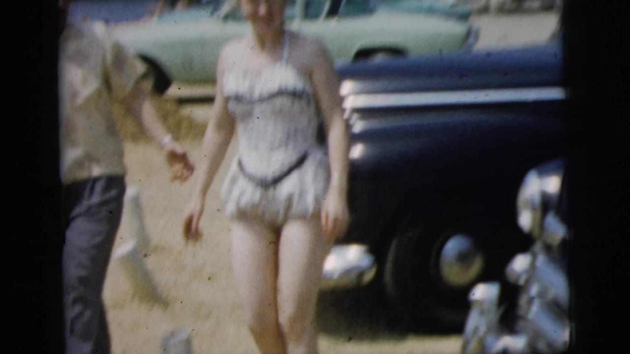 Woman in a swimsuit walks past parked cars while another person stands nearby.