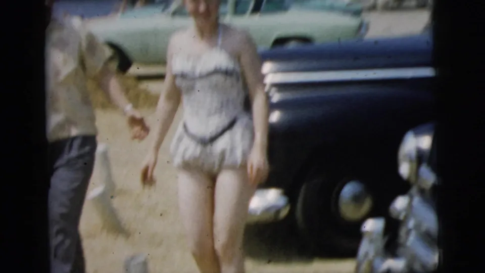 A woman in a light patterned swimsuit walks near dark and green parked cars in the 1961 Camden home-movie scene.