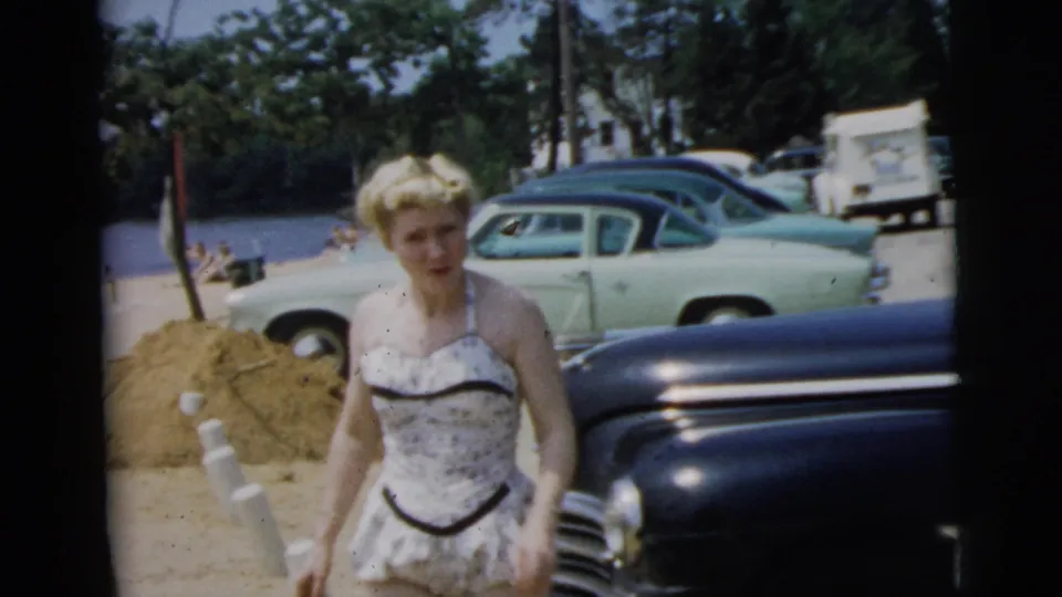 A blonde woman in a white patterned swimsuit stands near parked mid-century cars in Camden, New Jersey, according to the clip metadata.
