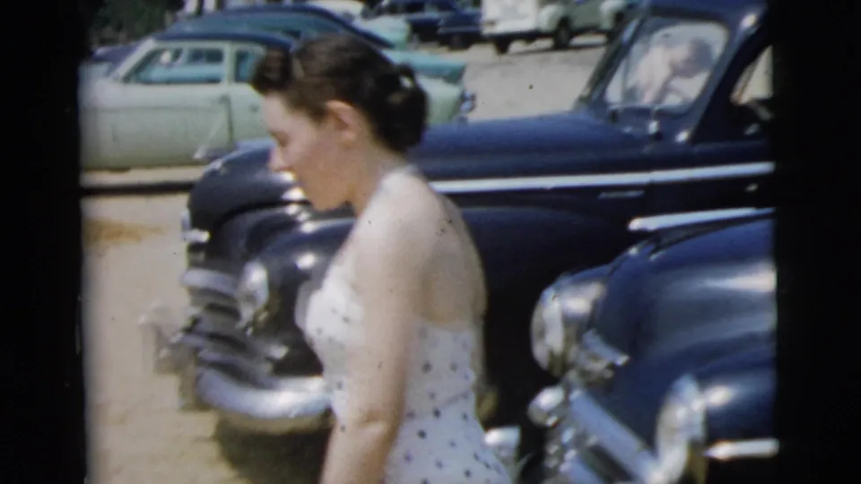 A side view shows a woman in a white patterned swimsuit walking beside dark parked cars in the Camden 1961 home-movie footage.