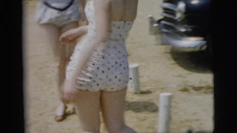 1961 Camden, New Jersey beach scene with a woman in a white polka-dot one-piece swimsuit walking past other people on sand.