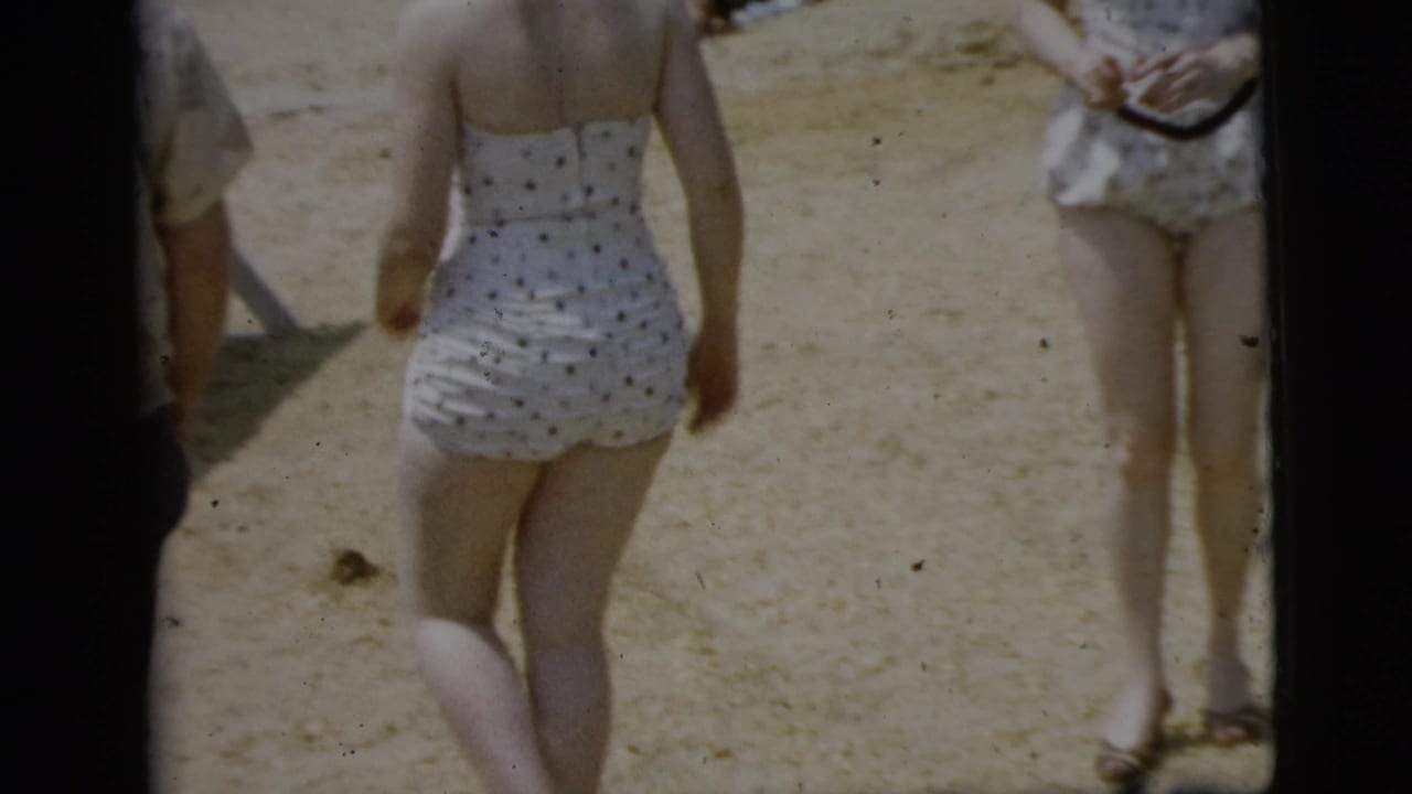 Woman in white swimwear seen from behind near a dark vintage car on sand.