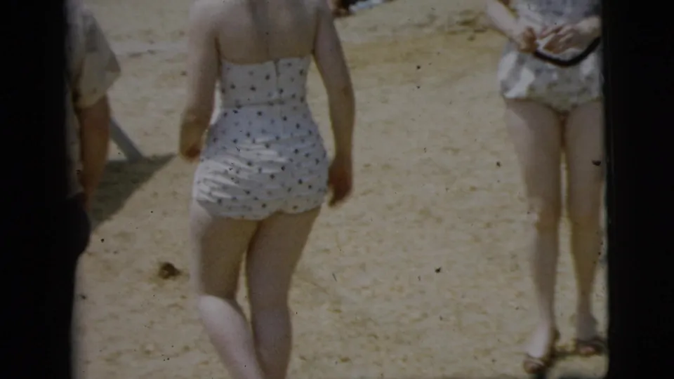 A woman in a white polka-dot swimsuit walks away from the camera on a Camden beach as a dark car passes or sits nearby.