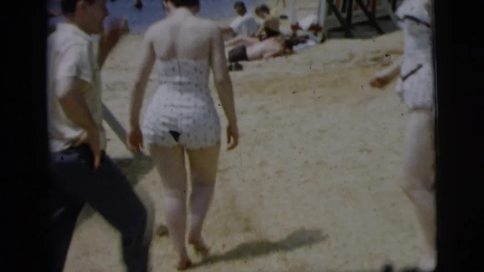 Home-movie view of a woman in a white polka-dot swimsuit walking through a busy Camden beach scene with people nearby.