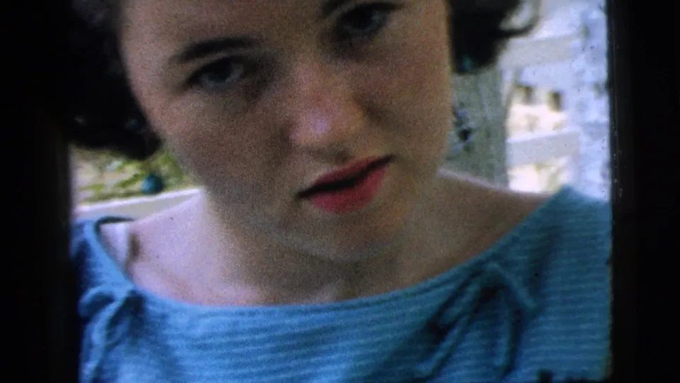 A girl in a blue dress looks toward the handheld camera outdoors, with a pale fence and yard details behind her.