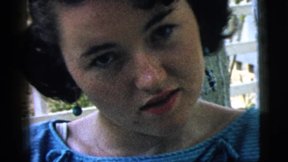 The girl in a blue dress is framed close to the camera near a white fence, with soft 8mm grain and handheld motion.