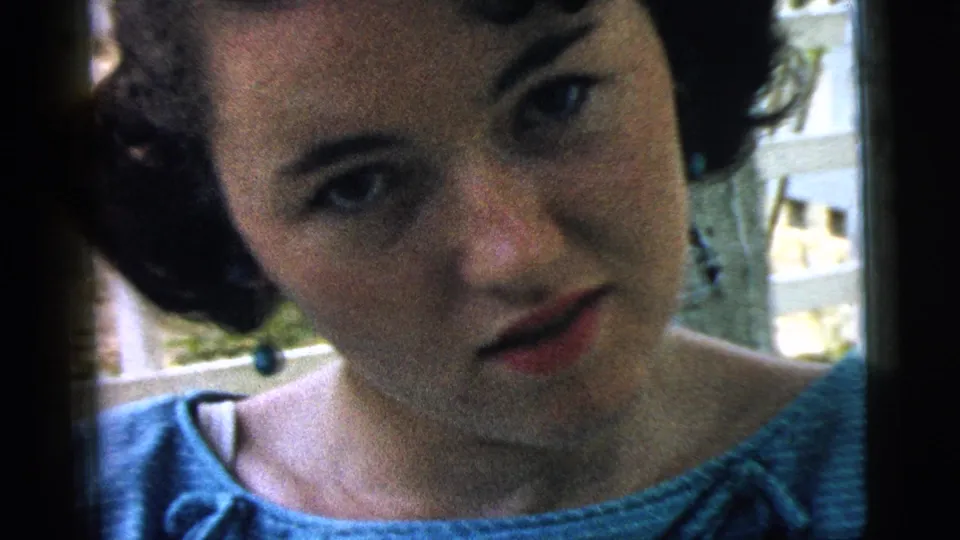 In 1961 Camden home-movie footage, a young woman in a blue top looks toward the camera near a white porch railing.