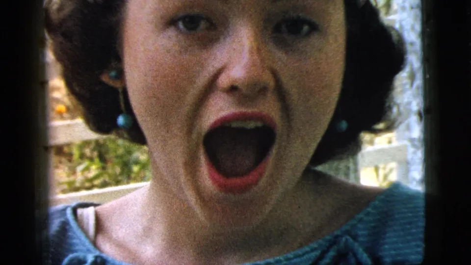 Young woman in Camden, New Jersey, laughs in close-up on 1961 8mm home-movie footage, with garden foliage behind her.