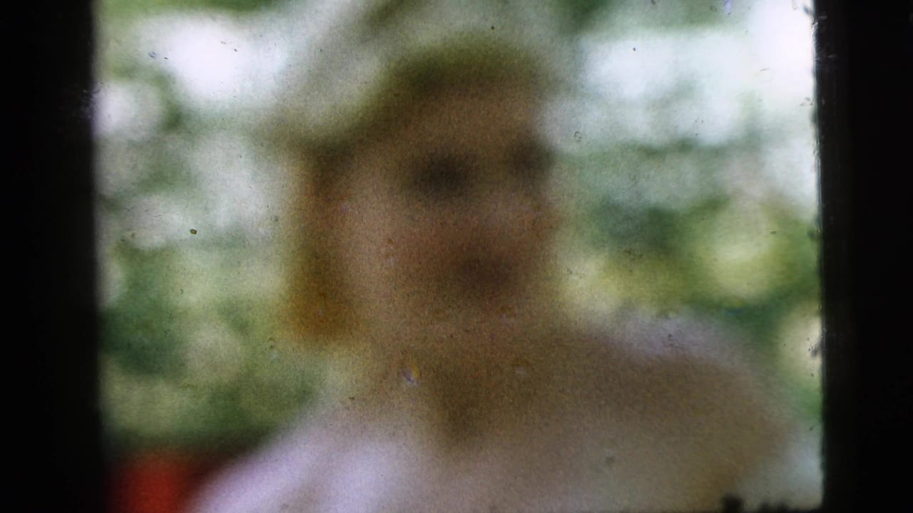 Blurred person in a light shirt seated outdoors with greenery behind.