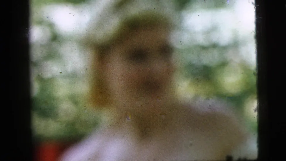 Soft-focus 1961 Camden home-movie frame showing a seated figure in a light shirt against green outdoor background.