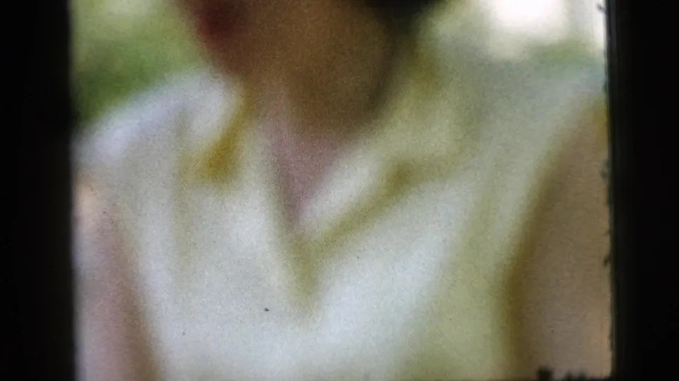 Blurred 1961 Camden frame of a woman in a pale blouse with yellow trim, seen close to the camera near a porch post.